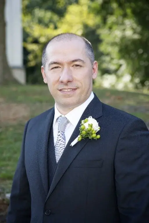 Ritratto di Matteo in abito scuro con cravatta e fiore bianco all'occhiello, nel verde